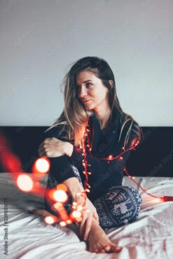 Fotografia Young Woman With Christmas Lights Sitting On Bed